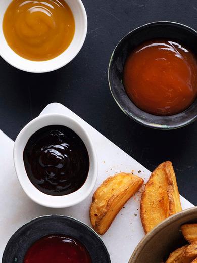Photo_On a black background, two pots of sauce of different flavours. Two more pots of BBQ sauce, slices of grilled potato, all on a white breadboard. Imagebild-Dipsaucen 2